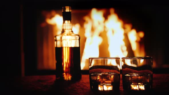 Two Glasses and a Bottle of Whiskey Stand on the Table Against the Background of a Hot Fireplace