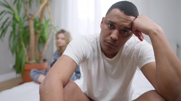 Portrait of Stressed Sad African American Young Man Looking at Camera Sitting on Bed with Blurred