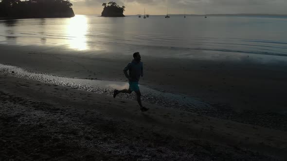 Aerial shot tracking a silhouetted young man running on a beach in Auckland, New Zealand