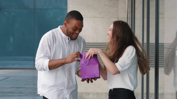 Multiracial Couple Standing Near Shop Building Outdoors Caucasian Woman Showing Gift Bag Purchase to