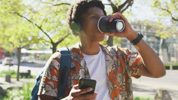 Happy african american man in city, using smartphone and headphones, drinking coffee in street