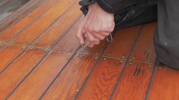 Side shot carpenter uses knife to re old mastic from roof planking of boat cabin