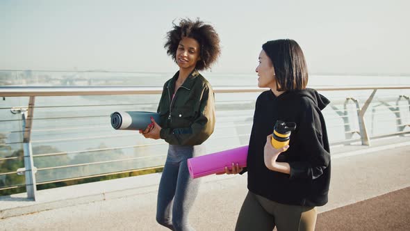 Happy Asian and African American Women Going to Yoga Class Talking on Walk Holding Fitness Mats Slow