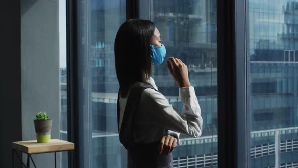 Thoughtful asian woman wearing face mask looking out of window at modern office