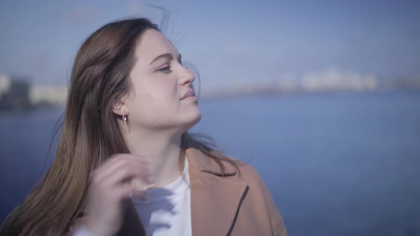 Close-up Face of Young Happy Caucasian Woman Sitting on River Bank and Fixing Hair. Cute Chubby Girl