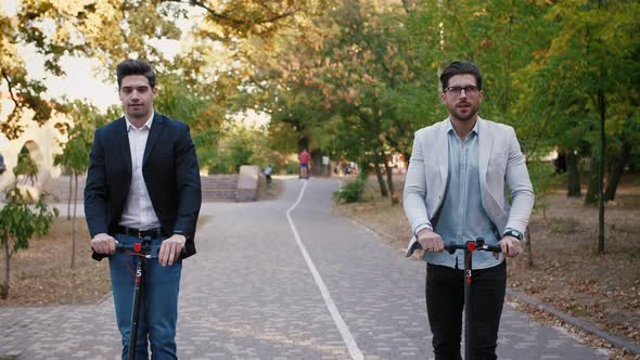 Portraits of Two Handsome Young Businessman Riding an Electric Scooters on the Road in City Park