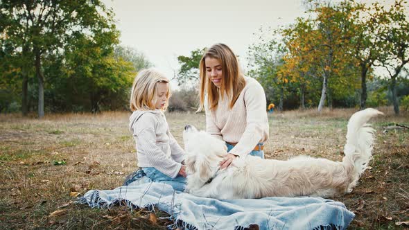 Young Mom and Little Daughter Sitting on Picnic Blanket at a Glade of Autumn Park Laughing and
