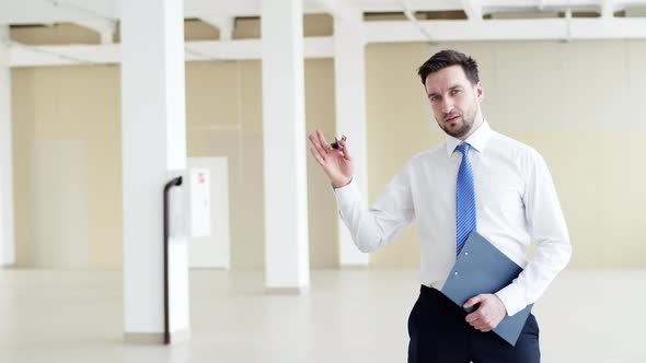 Male realtor stands in middle of rented office space and holds keys, front view. Realtor holds keys