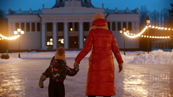 A Family of Mother and Two Kids Skating on Public Ice Rink at Evening in Bright Lights - Back View