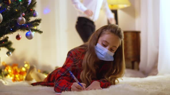 Concentrated Girl in Covid19 Face Mask Writing Letter to Santa Clause As Boy Entering Living Room