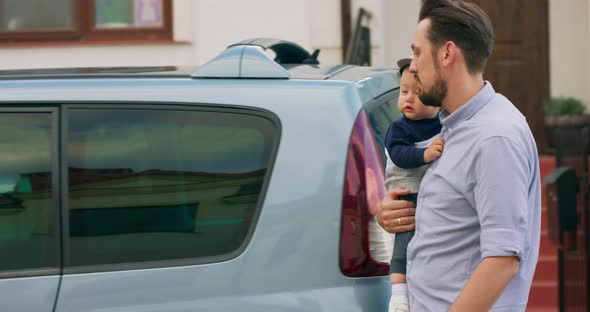 Young Bearded Father is Carrying Baby Boy and Puts Him Into the Baby Car Seat Inside of Family Car