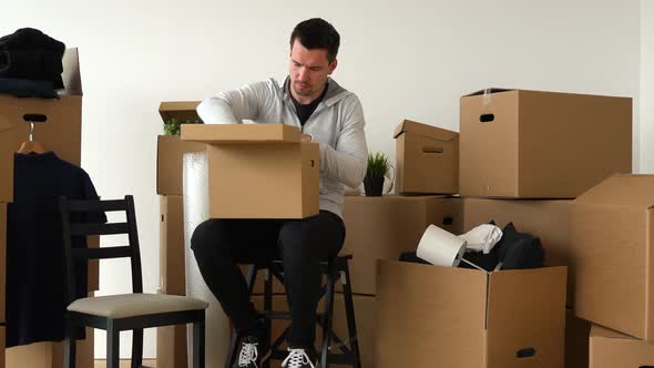 A Moving Man Sits on a Chair and Puts Things Into a Cardboard Box in an Empty Apartment