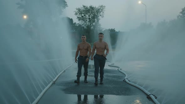 Two Muscular Men with Water Droplets on Their Naked Torsos Goes Among Water Jet From Spray Hoses