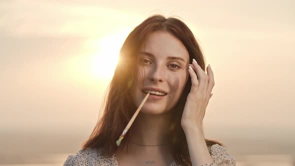 Young Pretty Woman Artist with Freckles on Her Face and with Ginger Hair Bites Painting Brush and