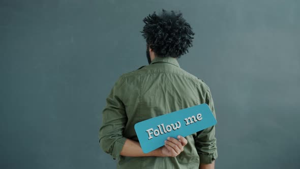 Goodlooking Arab Man Standing with Back to Camera Holding Follow Me Sign and Turning Smiling