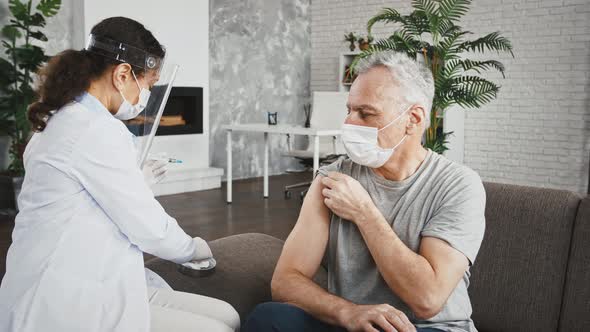 Woman Doctor in Protective Mask and Face Shield is Injecting Vaccine Into Shoulder of Aged Man Who