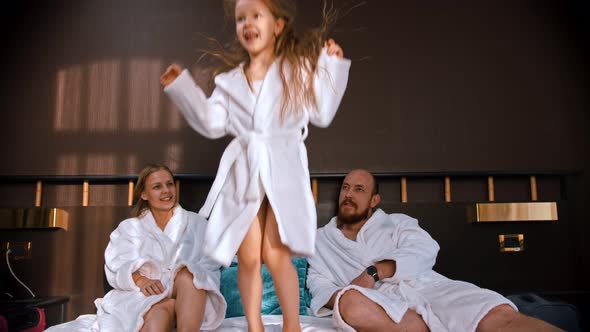 A Family in White Bathrobes Sitting in the Bed - a Girl Jumping on the Bed and Her Mother Catches