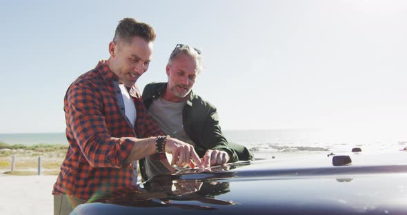 Happy caucasian gay male couple leaning on car, reading map and talking on sunny day at the beach