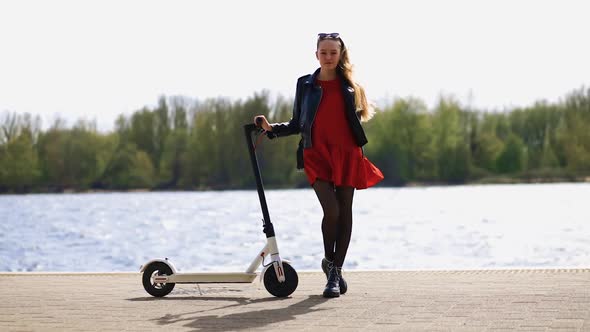 Young Stylish Woman Stands and Holds Electric Scooter on Promenade