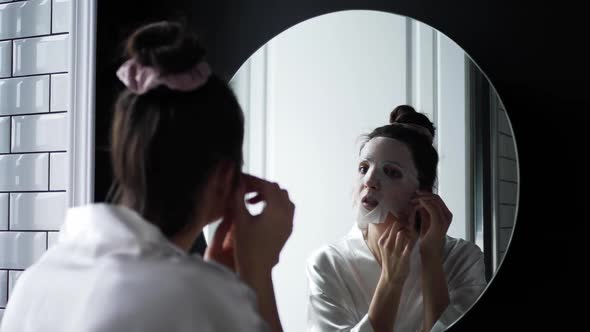 Woman in the Bathroom in Front of the Mirror Puts on a Rejuvenating Face Mask From Wrinkles