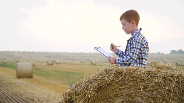 Little Farmer on Top of a Straw Bale Holds a Tablet with a Letter and a Pen in His Hands on a