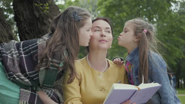 Mature Woman Hugging Her Two Cute Granddaughters Sitting on the Grass Under the Tree in the Park