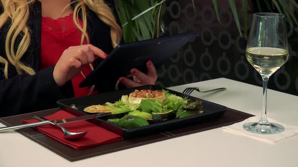A Woman Sits at a Table in a Restaurant and Works on a Tablet, a Salad in Front of Her