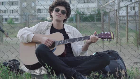 Portrait of Romantic Thoughtful Musician Playing Guitar Outdoors As Other Guys Playing Football at