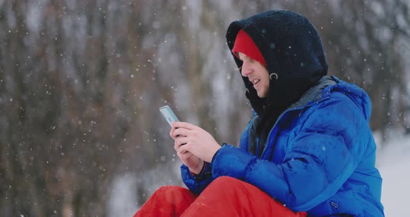 A Male Snowboarder Sitting on the Snow Takes Photos on the Phone of a Beautiful Resort Landscape