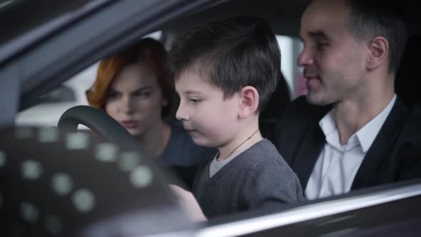 Close-up Portrait of Cute Little Caucasian Boy Sitting in Car with Parents and Holding Steering