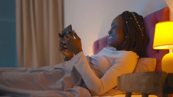 Beautiful Young African Woman Wearing Pajamas, Lying in Bed Checking Email on Tablet Computer