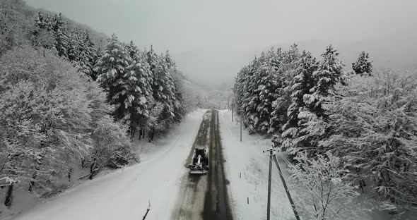 Aerial view of snowy forest and highway while snow cleaner car removes snow from the road