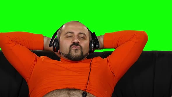 Funny Fat Caucasian Guy Dancing When Sitting on Sofa in Big Headphones. Green Screen. Slow Motion