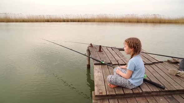 a Little Girl Sits with a Fishing Rod on a Bridge By the River