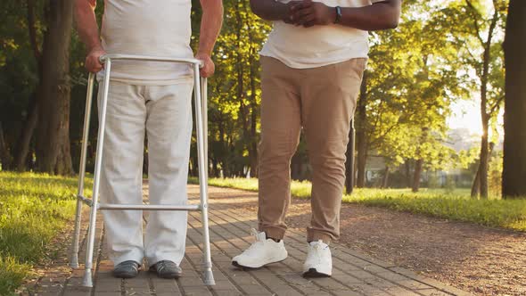 African-American caregiver is teaching disabled old man to walk with walker. Nurse and patient.