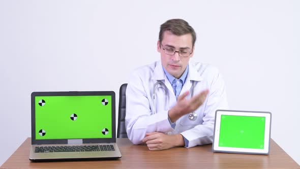 Young Handsome Man Doctor Presenting Something on Laptop and Digital Tablet Against Wooden Table