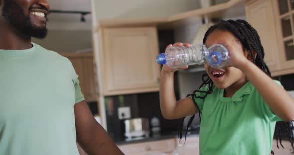 Happy african american father and daughter playing in kitchen with plastic bottles for recycling