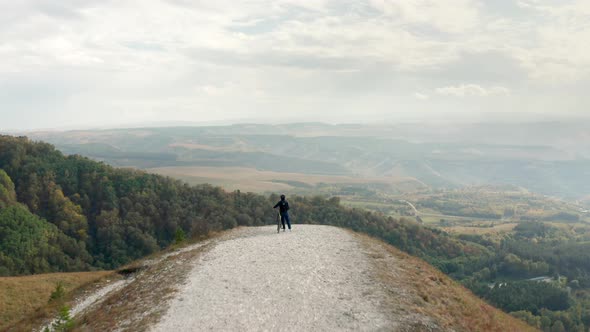 Biker in Helmet with Mtb Bicycle Stands on Hill Edge Looking at Forest in Valley Under Cloudy Sky on