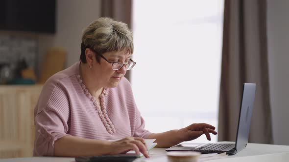 Old Woman is Learning Online Looking for Information in Internet By Laptop and Making Notes in