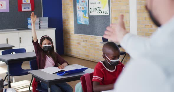 Diverse schoolchildren sitting in classroom raising hands during lesson, all wearing face masks