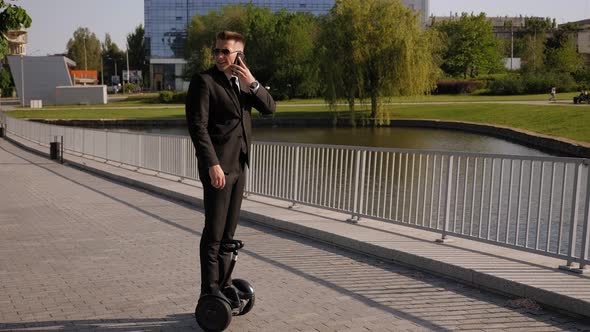 A Successful Businessman Rides a Gyro Scooter in a Park and Talks on the Phone