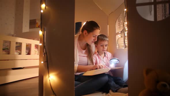 Panning Shot of Young Smiling Woman Reading Story Book To Her Little Son at Night While Sitting on