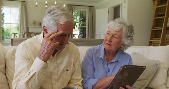 Stressed caucasian senior couple smiling while using digital tablet sitting on the couch at home