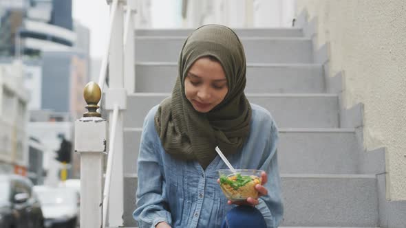 Woman wearing hijab with a takeaway salad using her phone sitting on stairs