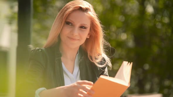 Young Beautiful Blonde Girl in Summer Day is Reading a Book Sit on a Bech at Urban City Park