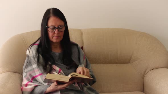Middle-aged woman seated in double armchair going through and reading The Bible.