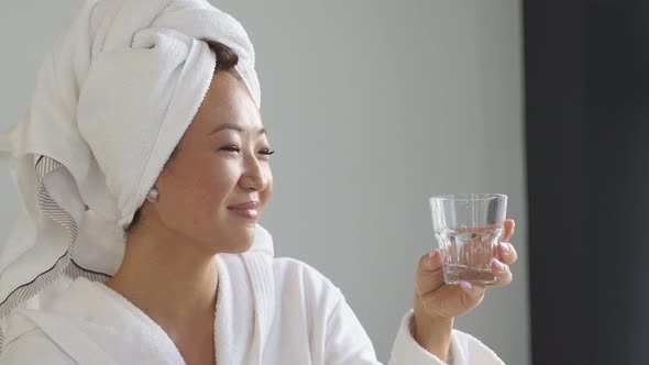 Portrait of a Young Woman Wearing a Bathrobe and a Towel on Her Head Drinking Water From a Glass