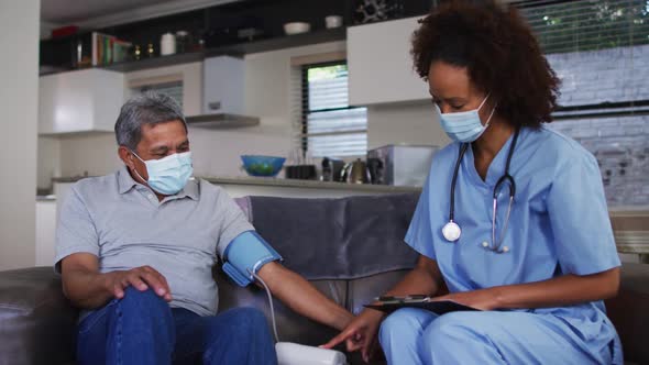 Mixed race female doctor wearing mask and senior man taking blood pressure at home