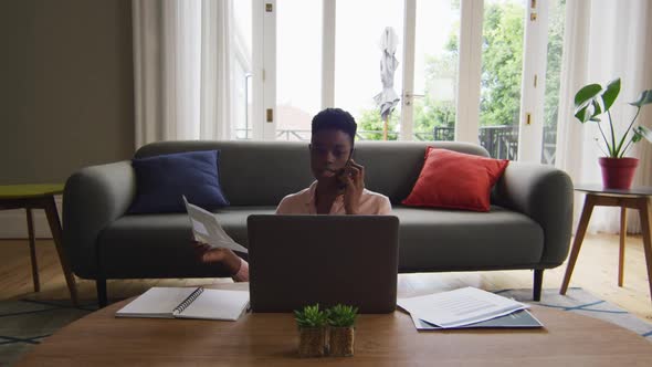 African american woman holding a document talking on smartphone while working from home