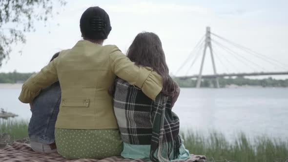 Back View of an Elegant Mature Woman Sitting on the Grass on the Riverbank, Hugging Granddaughters